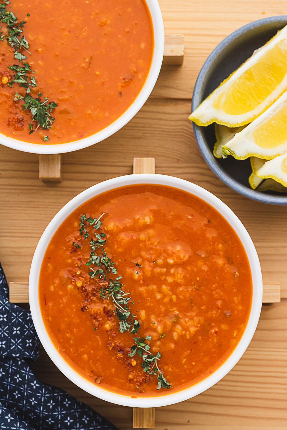 Two white bowls filled with fiery red Turkish Lentil Soup garnished with dried mint and red pepper flakes surrounded by lemon wedges and bread.