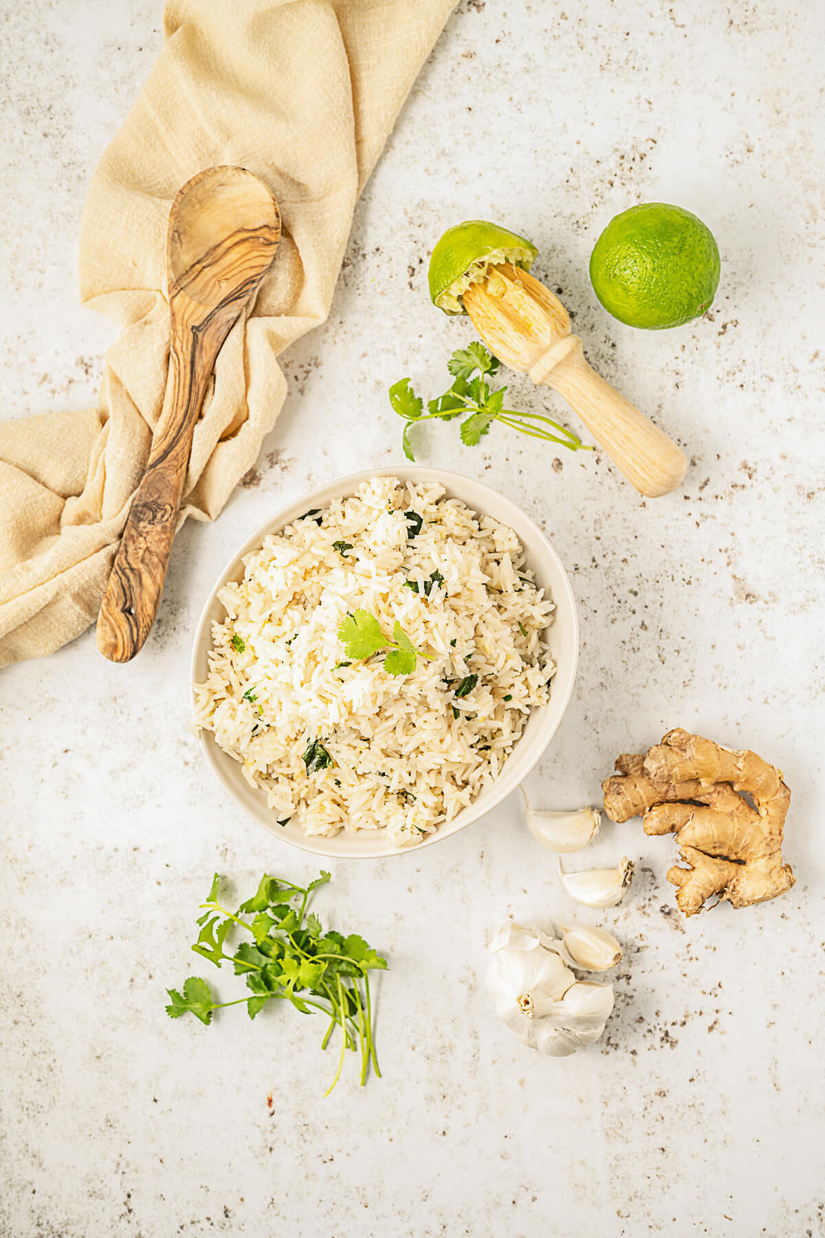 A white bowl full of Ginger Rice surrounded by fresh cilantro, limes, ginger root, and garlic cloves.