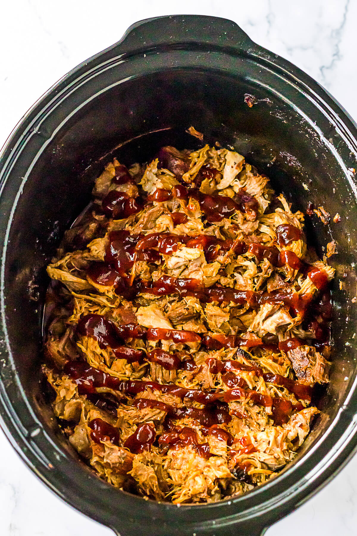 A black slow cooker insert full of pulled pork drizzled with barbecue sauce.