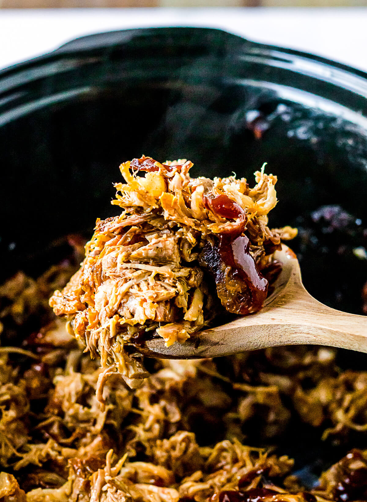 A wooden spoon holds a spoonful of shredded pork above a slow cooker filled with Root beer pulled pork.