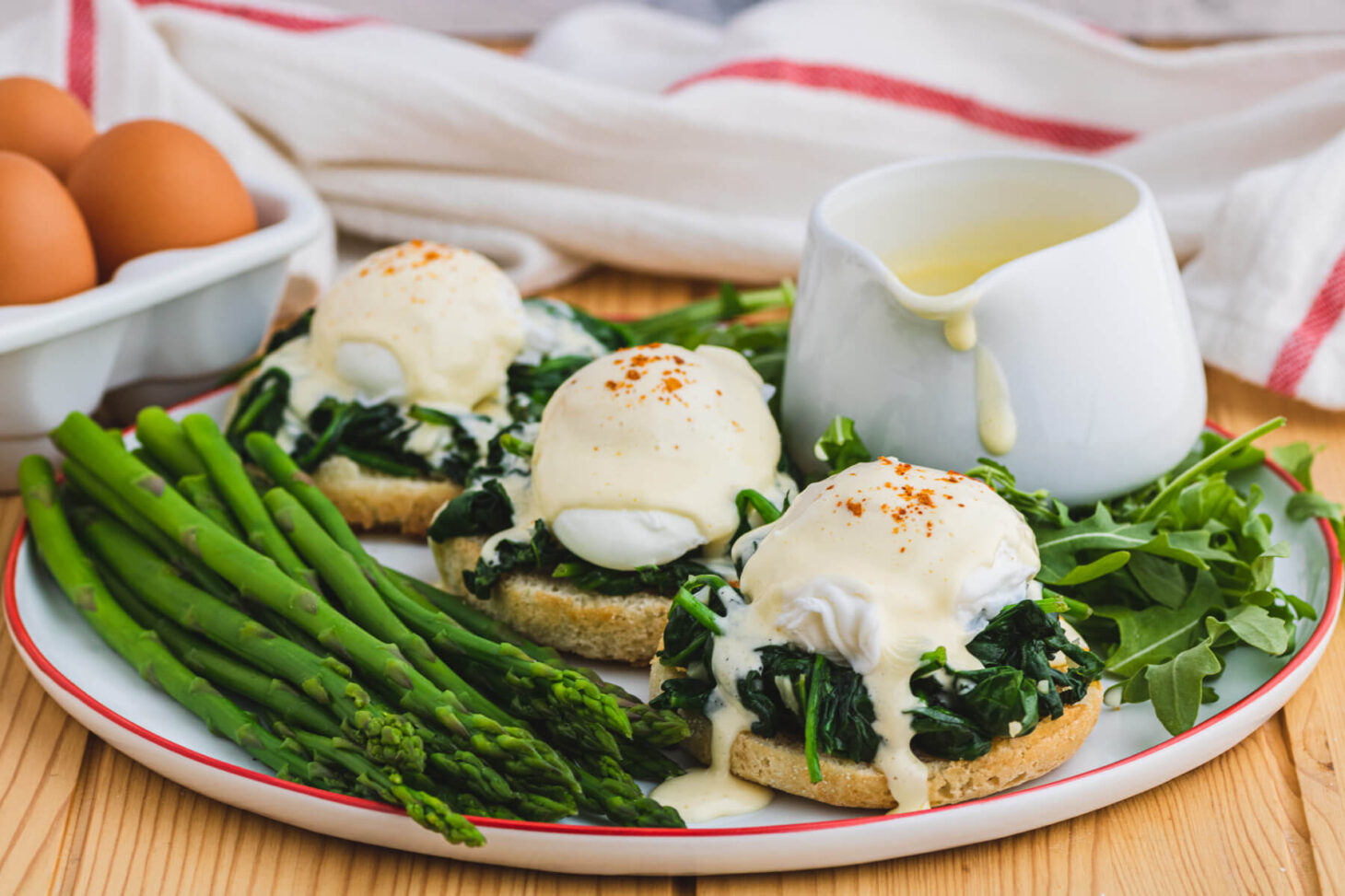 A red rimmed white plate holding two poached eggs covered in creamy yellow hollandaise sauce over a bed of spinach on toasted English muffins with a side of asparagus and baby arugula.