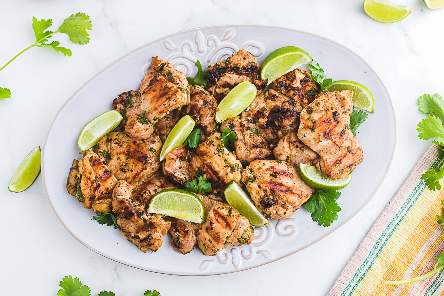 A white platter of grilled chicken thighs topped with chopped fresh cilantro and lime wedges.