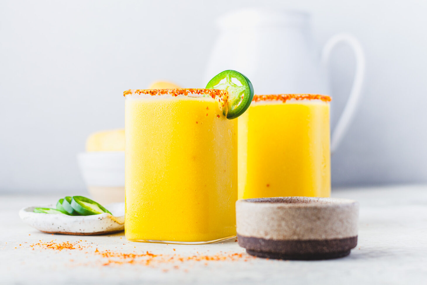 Two square rocks glasses filled with with vibrant spicy mango margarita garnished with jalapeños and Tajin seasoning.