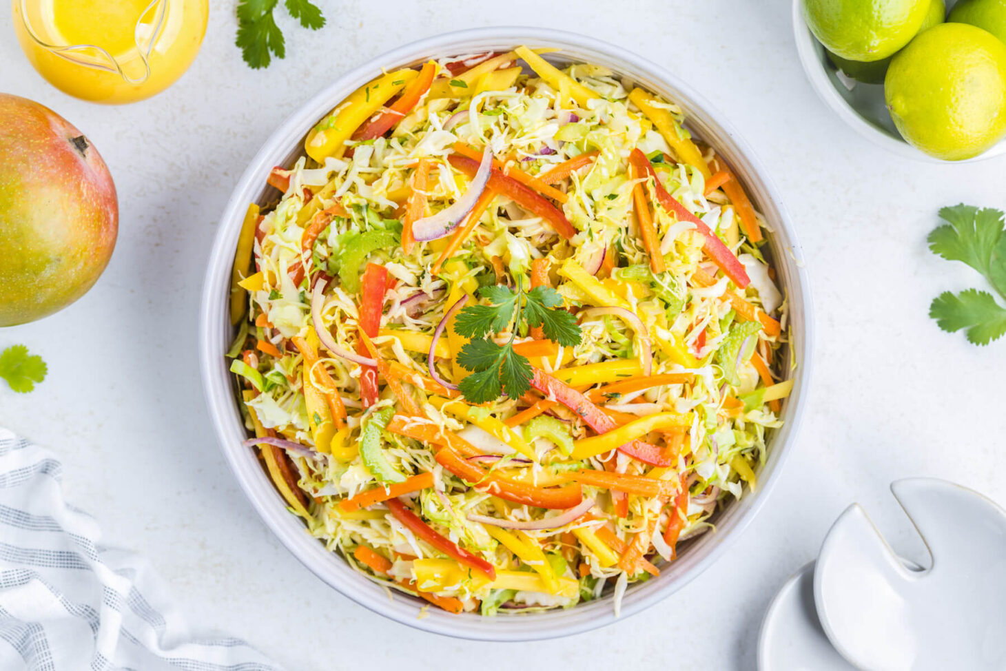 A white bowl filled with colourful mango slaw on a white background surrounded by mangos, limes, and cilantro.