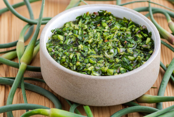 A small bowl containing vibrant green garlic scape chimichurri on a wooden table surrounded by fresh garlic scapes.