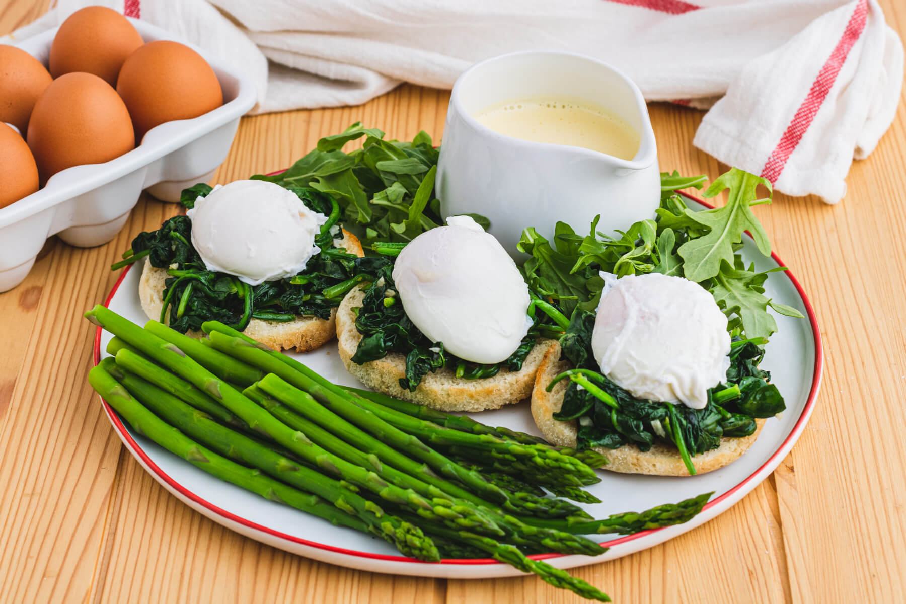 A red rimmed white plate holding two poached eggs ready to be covered in creamy yellow hollandaise sauce over a bed of spinach on toasted English muffins with a side of asparagus and baby arugula.