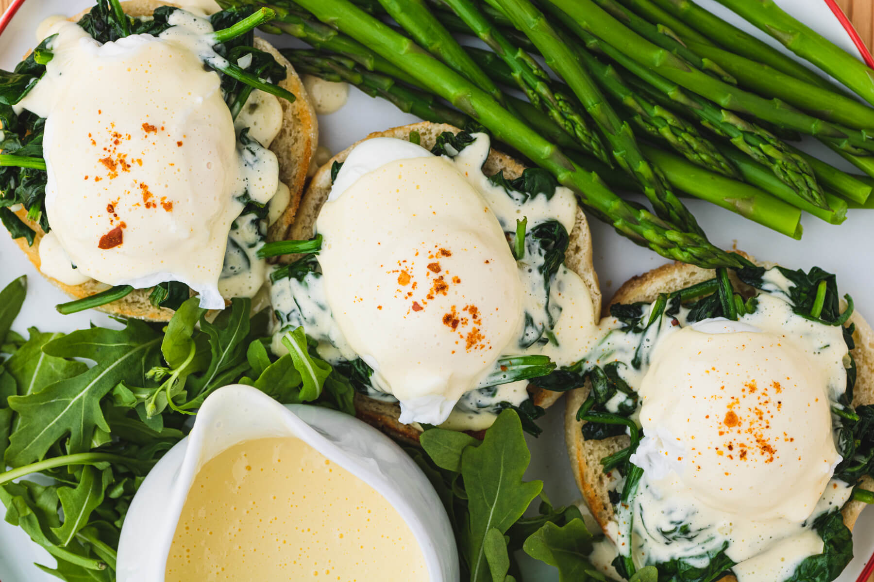 Three Eggs Benedict Florentines surrounded by verdant green asparagus and baby arugula with a pitcher of creamy yellow hollandaise.