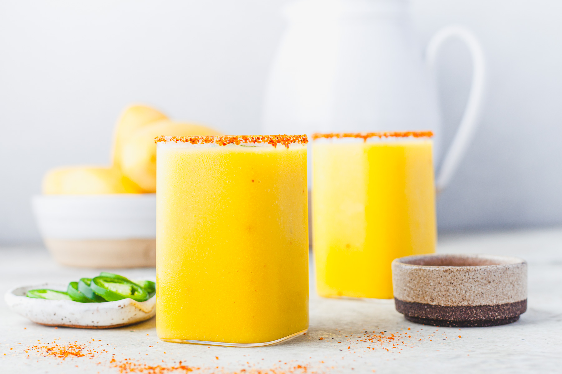 Two square rocks glasses filled with with vibrant spicy mango margarita garnished with Tajin seasoning.