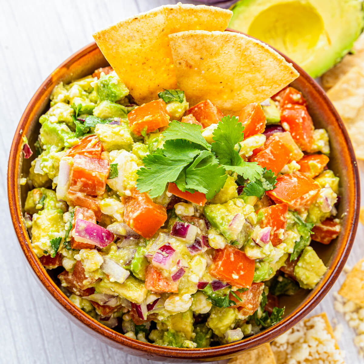 A terra cotta bowl full of chunky avocado salsa featuring avocado, tomatoes, cilantro, Cotija cheese, and red onions garnished with tortilla chips.