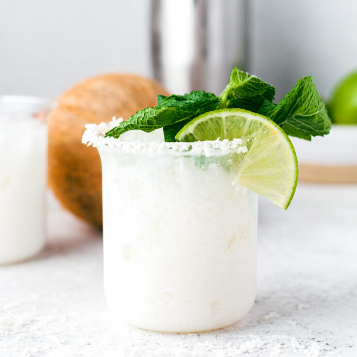 A creamy coconut margarita in a rocks glass rimmed with coconut and garnished with fresh mint and lime wedges.