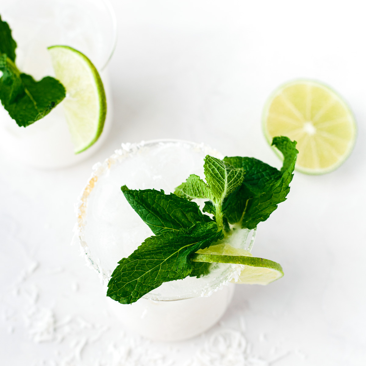 Two creamy coconut margaritas in rocks glasses rimmed with coconut and garnished with fresh mint and lime wedges.