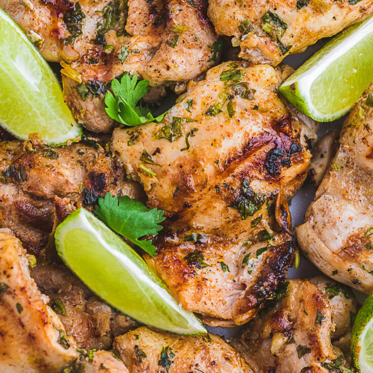A plate of grilled chicken thighs topped with chopped fresh cilantro and lime wedges.