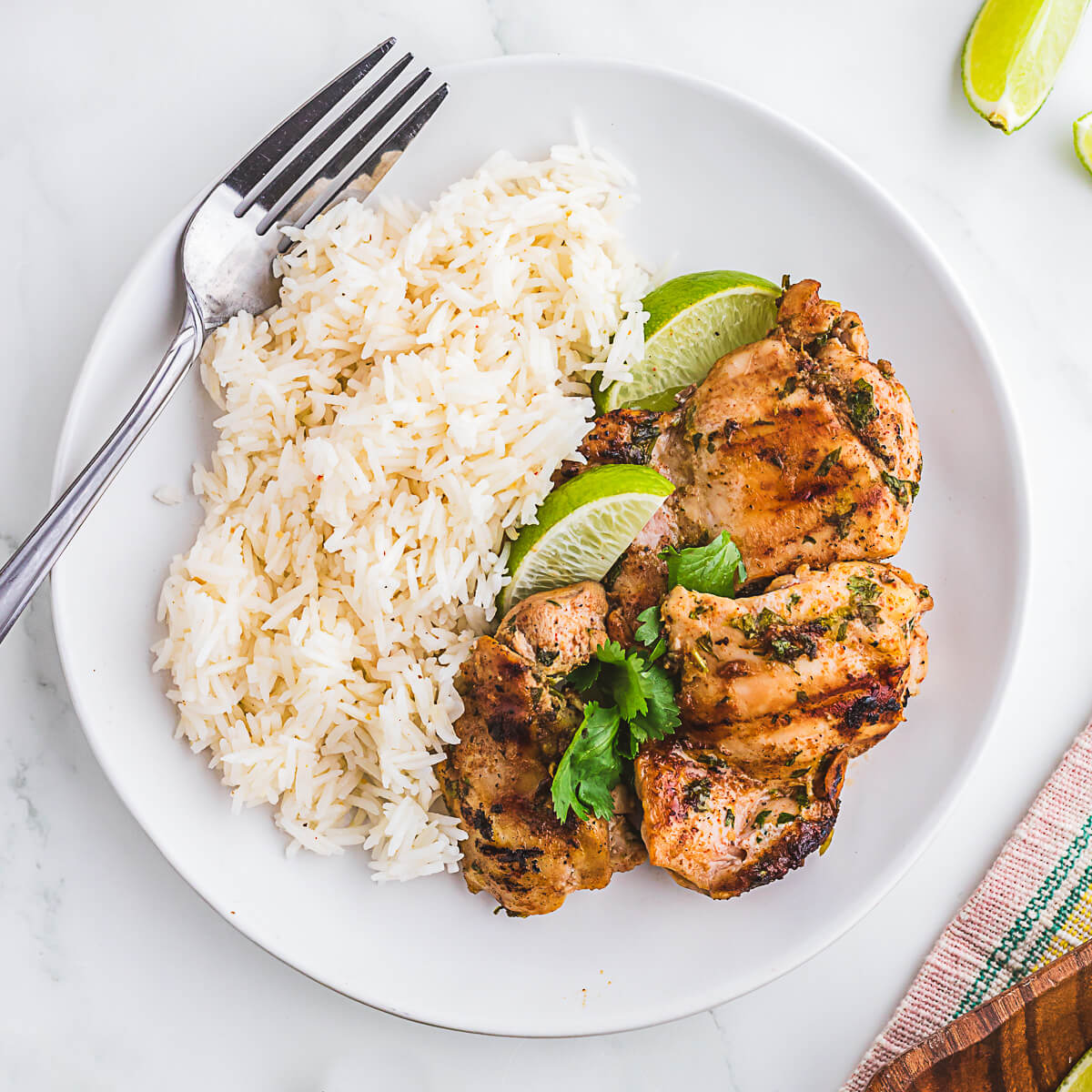 A plate of grilled chicken thighs garnished with cilantro and served with a side of white rice and lime wedges.