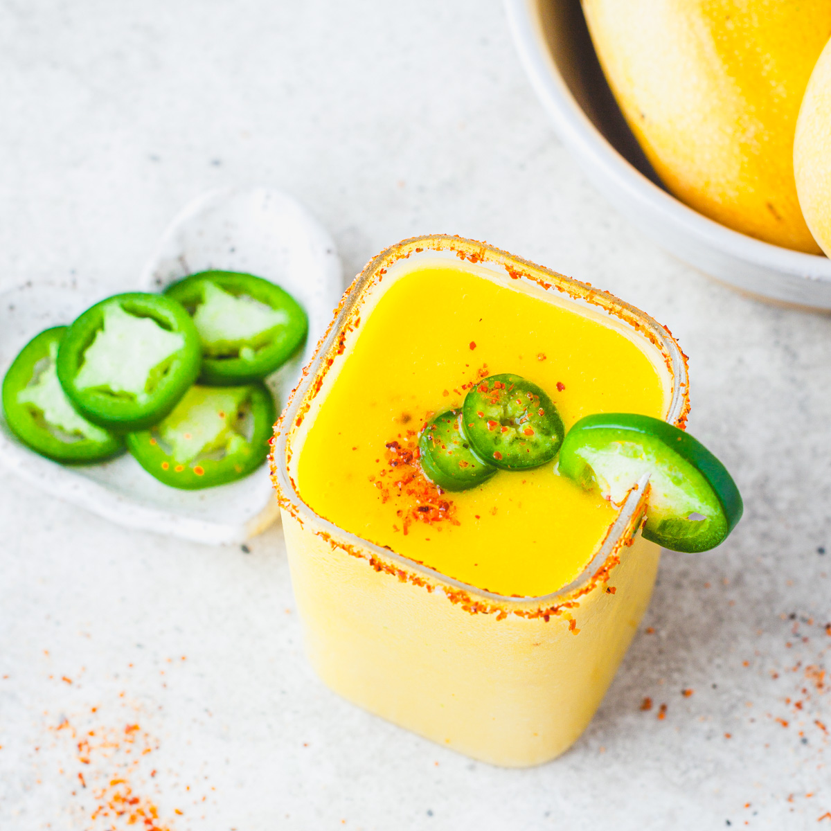 A square rocks glass filled with with vibrant spicy mango margarita garnished with jalapeños and Tajin seasoning beside a bowl of mangos.