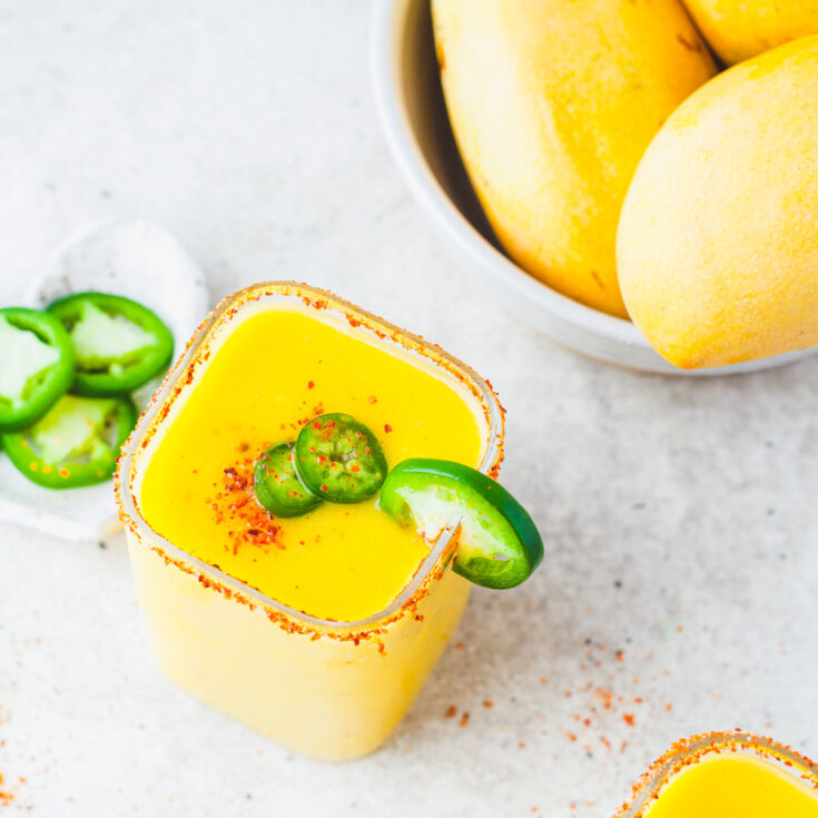 A square rocks glass filled with with vibrant spicy mango margarita garnished with jalapeños and Tajin seasoning beside a bowl of mangos.