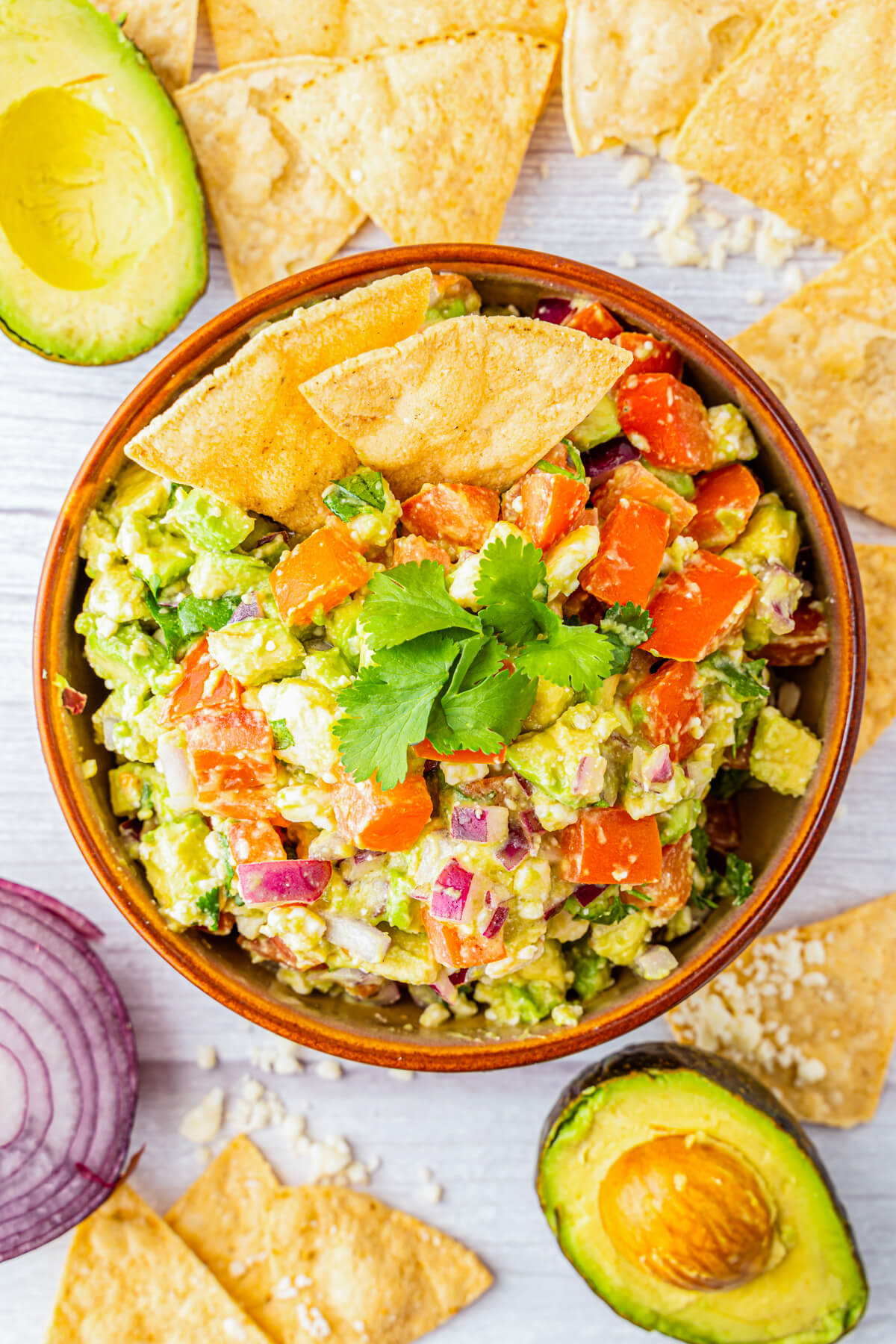 A terra cotta bowl full of chunky avocado salsa featuring avocado, tomatoes, cilantro, Cotija cheese, and red onions garnished with tortilla chips.