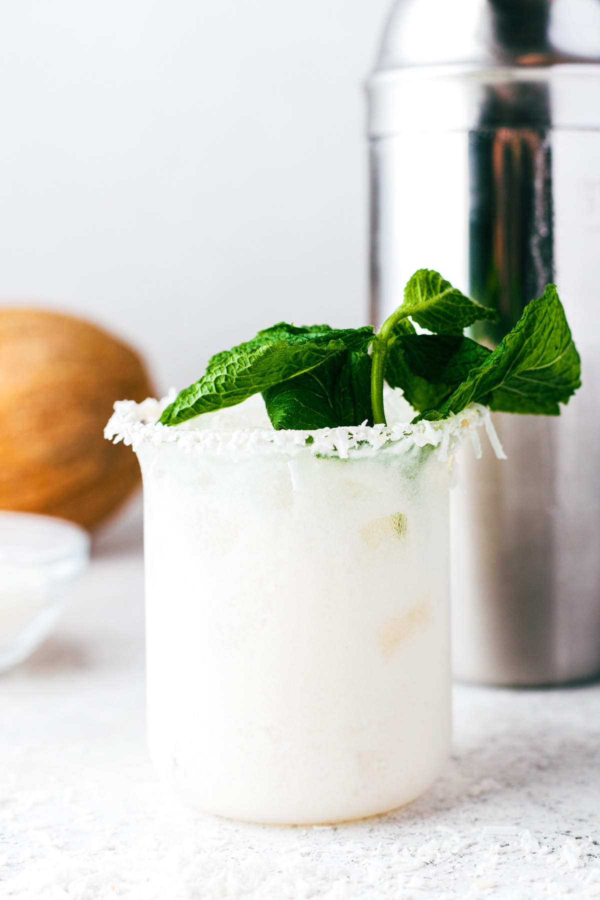 A creamy coconut margarita in a rocks glass rimmed with coconut and garnished with fresh mint.