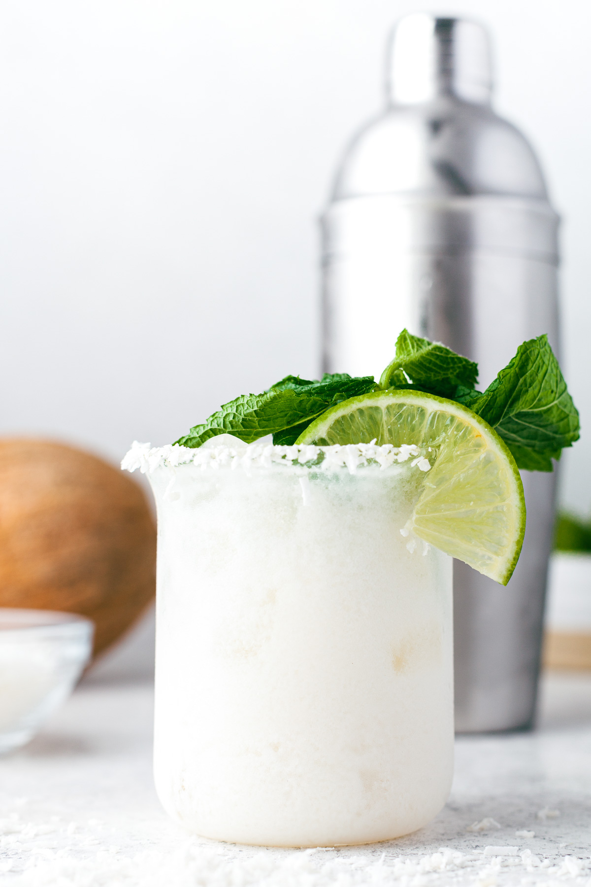A creamy coconut margarita in a rocks glass rimmed with coconut and garnished with fresh mint and lime wedges.