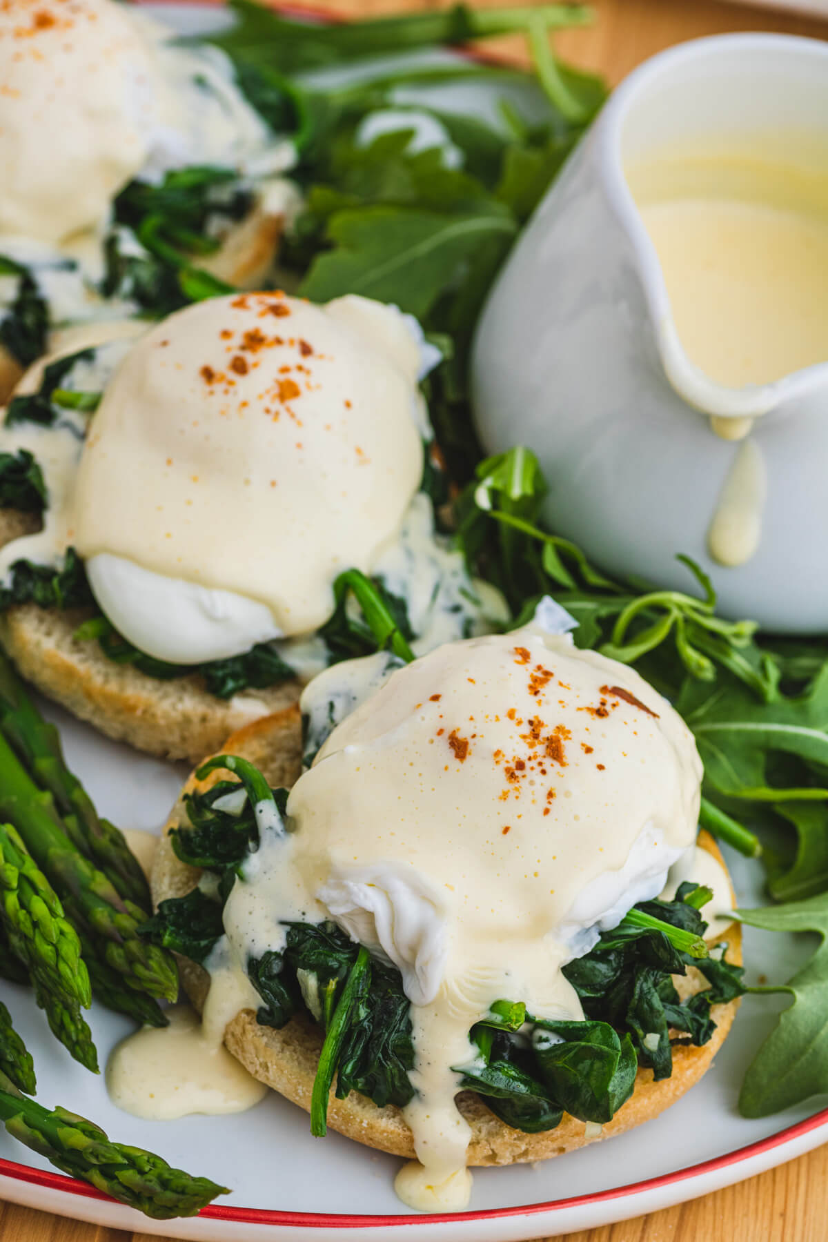 A red rimmed white plate holding two poached eggs covered in creamy yellow hollandaise sauce over a bed of spinach on toasted English muffins with a side of asparagus and baby arugula. 