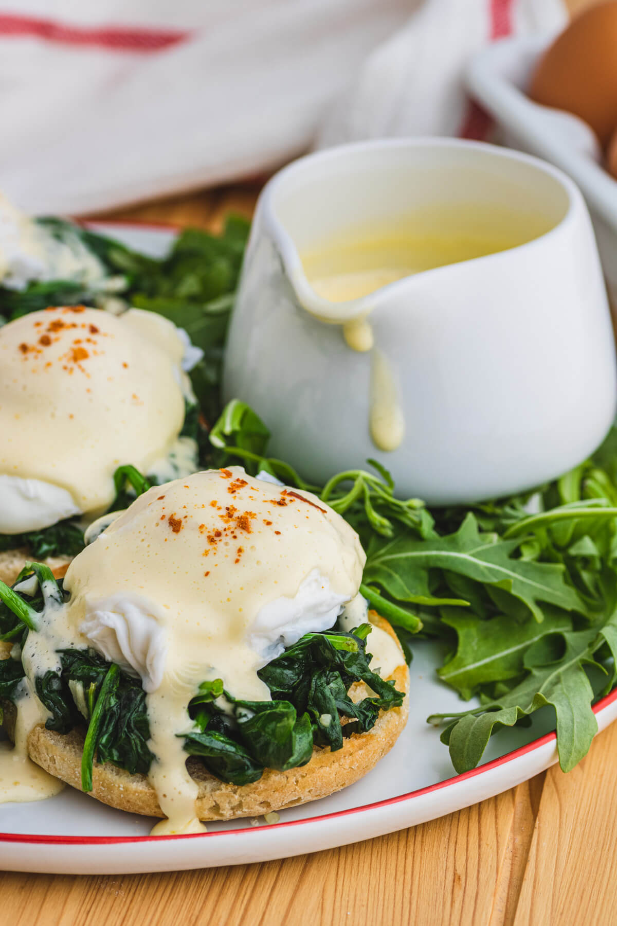 A red rimmed white plate holding two poached eggs covered in creamy yellow hollandaise sauce over a bed of spinach on toasted English muffins with a side of asparagus and baby arugula.