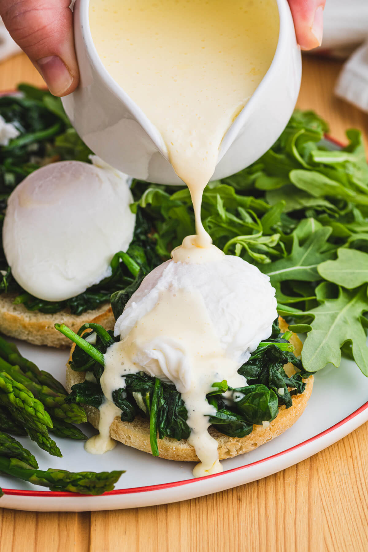 A hand pours creamy yellow hollandaise sauce over a poached egg laying on a bed of sauteed spinach and English muffin. 