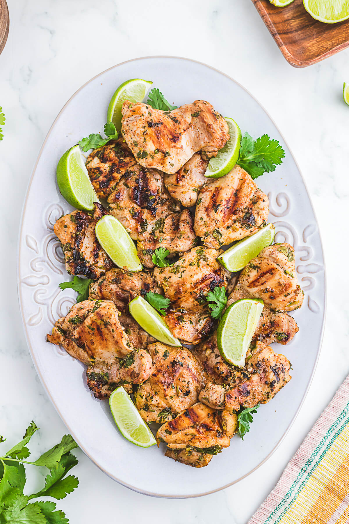 A white platter of grilled chicken thighs topped with chopped fresh cilantro and lime wedges.