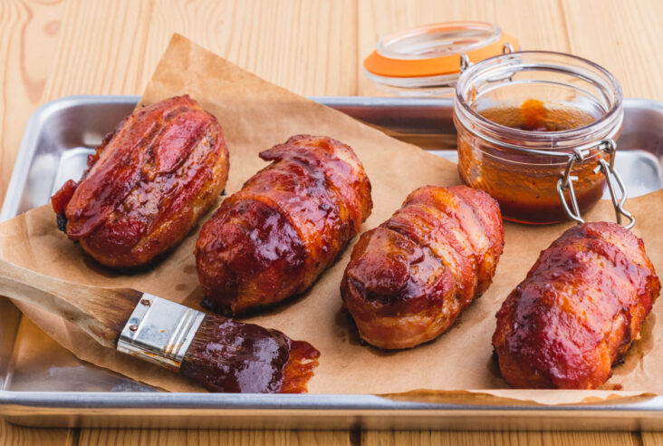 Four bacon wrapped and glazed Armadillo Egg appetizers on butcher paper beside a jar of BBQ sauce and brush full of the same BBQ sauce.