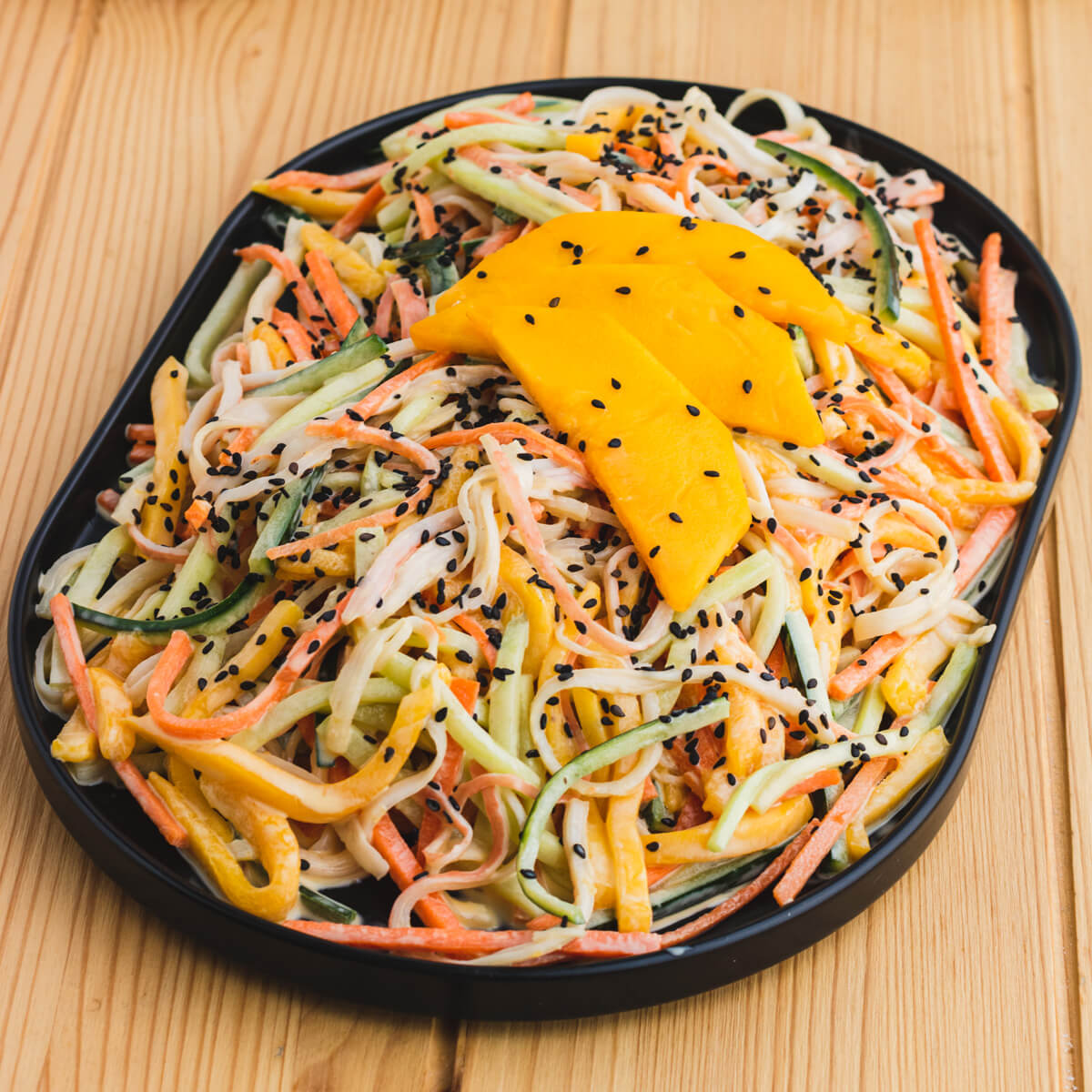 A black platter filled with shredded Kani salad topped with slices of mango, black sesame seeds, and chopped scallions.