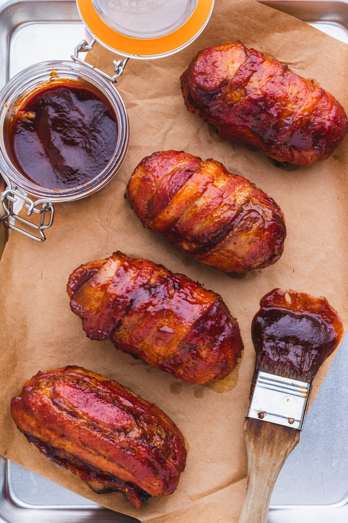 Four bacon wrapped and glazed Armadillo Egg appetizers on butcher paper beside a jar of BBQ sauce and brush full of the same BBQ sauce.
