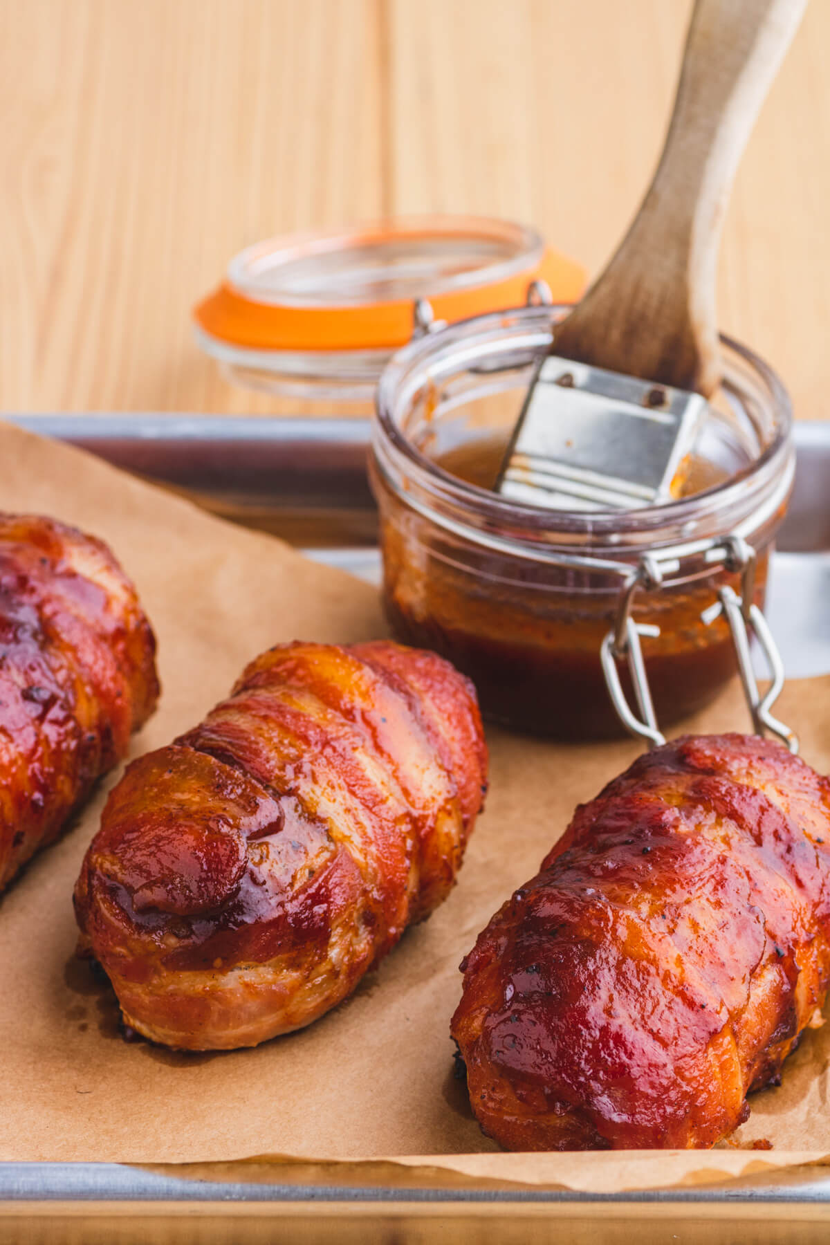 Three bacon wrapped and glazed Armadillo Egg appetizers on butcher paper beside a jar of BBQ sauce and brush full of the same BBQ sauce.