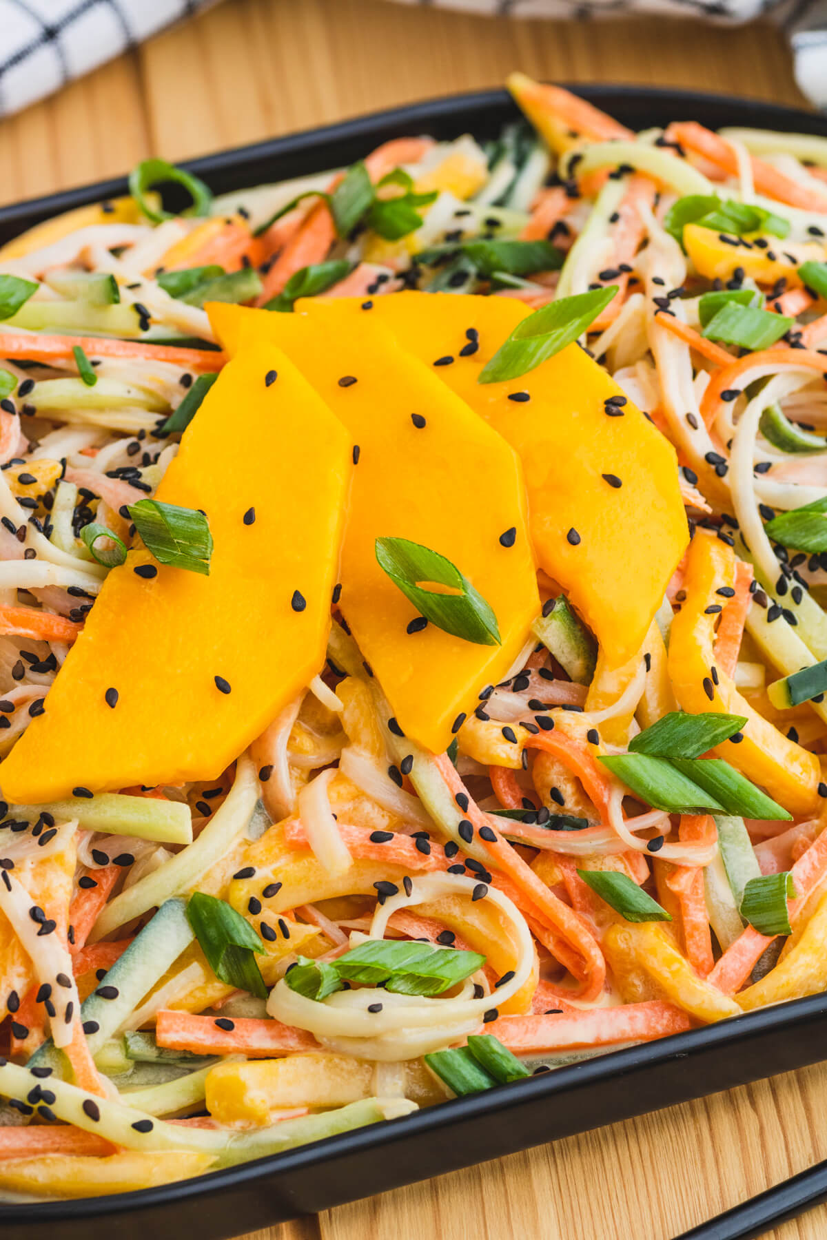 A black platter filled with shredded Kani salad topped with slices of mango, black sesame seeds, and chopped scallions.