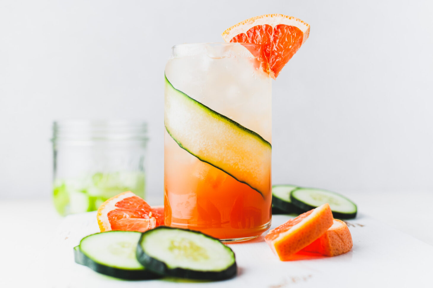 A cocktail glass lined with a thin slice of cucumber and filled with ice and pink grapefruit cocktail with a small wedge of grapefruit as a garnish.