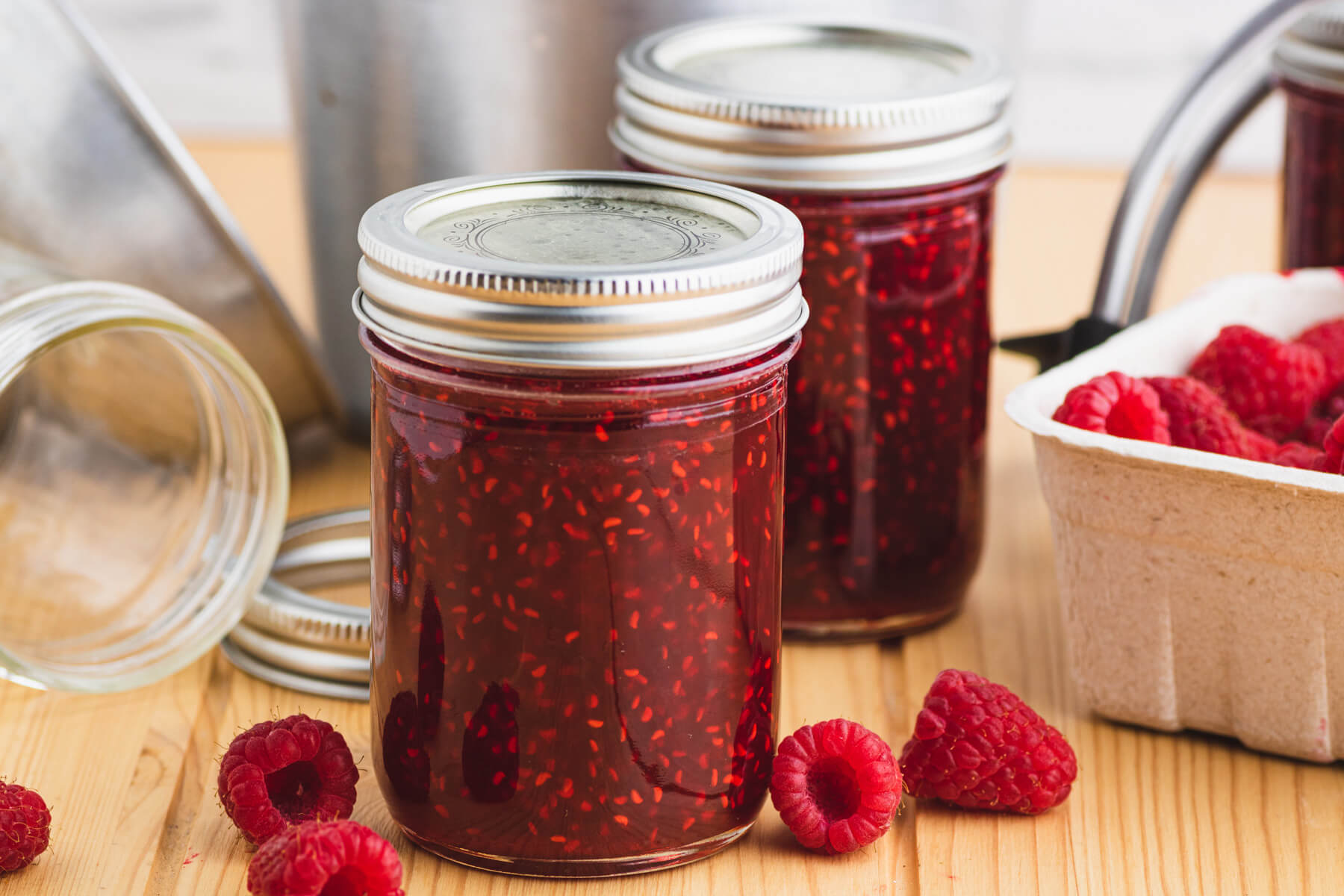 Two jars of vibrant red raspberry jam surrounded by tools and ingredients needed to make the recipe.