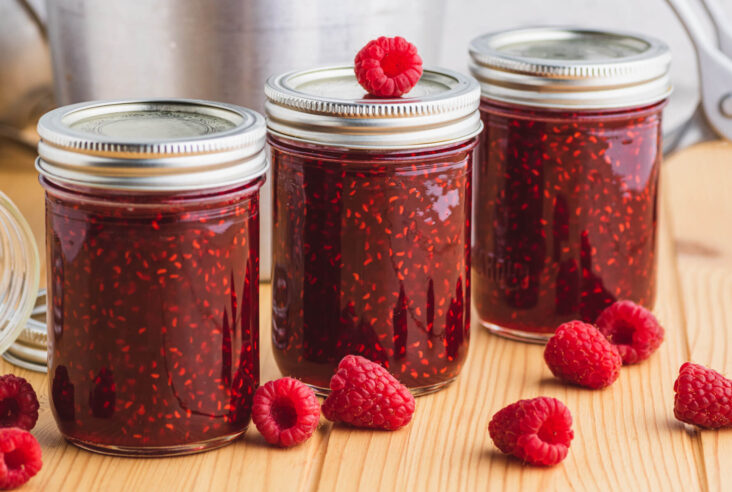 A fresh raspberry sits on top of one jar of raspberry jam in a group of three jars.