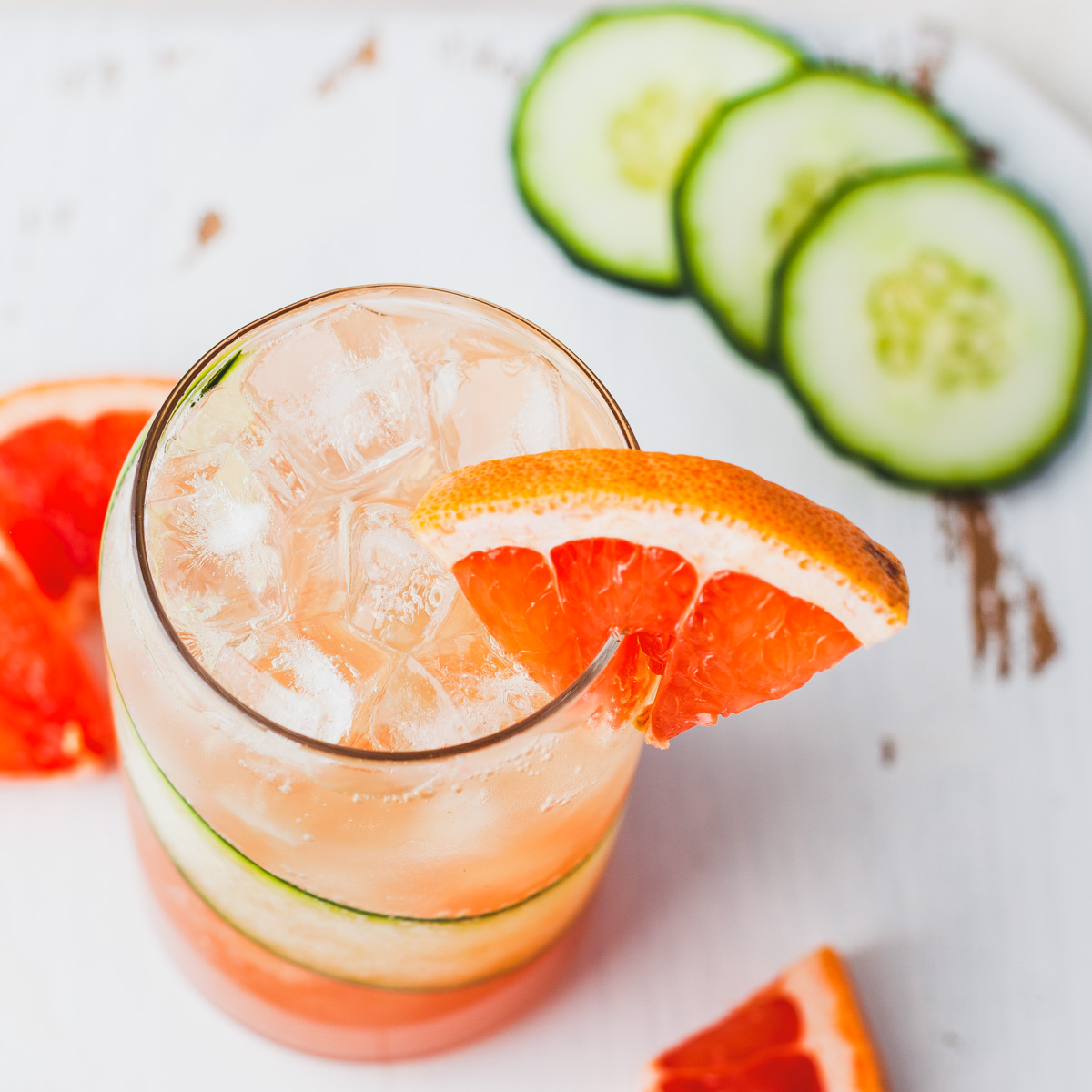 A cocktail glass lined with a thin slice of cucumber and filled with ice and pink grapefruit cocktail with a small wedge of grapefruit as a garnish.