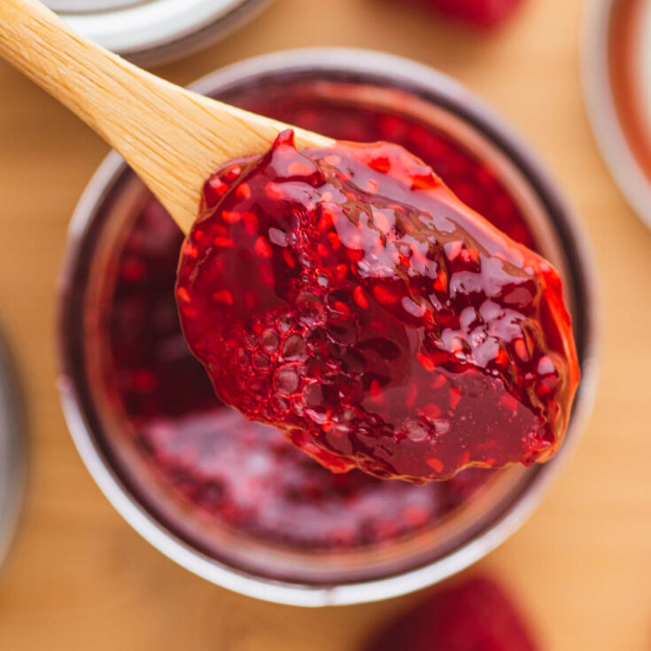 A wooden spoon holds a spoonful of raspberry jam over a jar of the same jam.