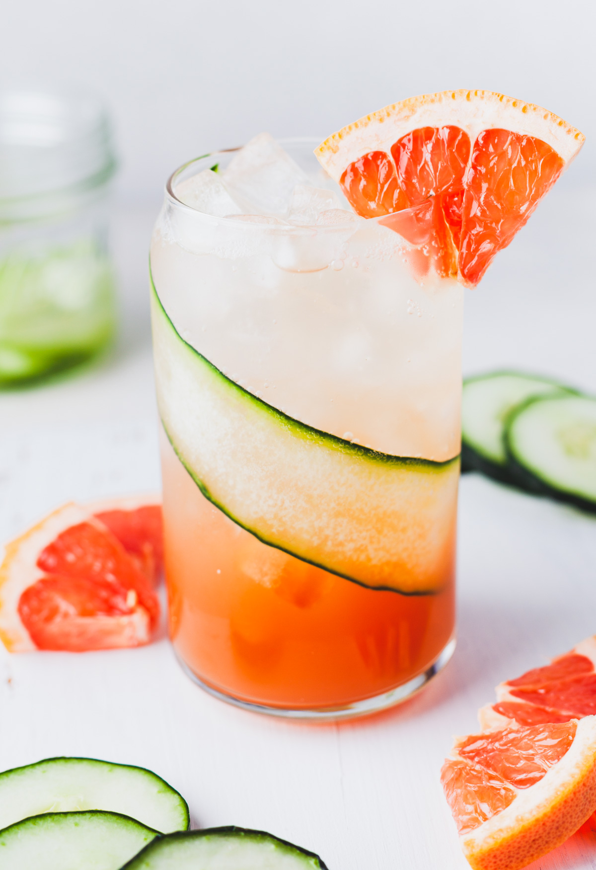 A cocktail glass lined with a thin slice of cucumber and filled with ice and pink grapefruit cocktail with a small wedge of grapefruit as a garnish. 