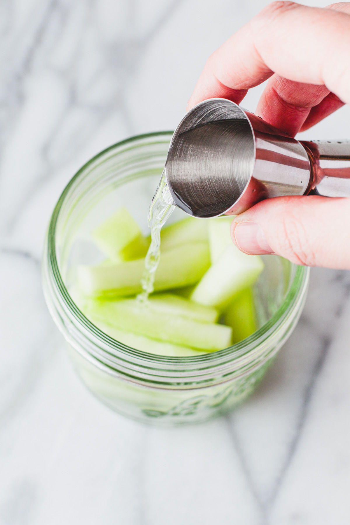 Gin being poured into a glass jar filled with sliced cucumber. 