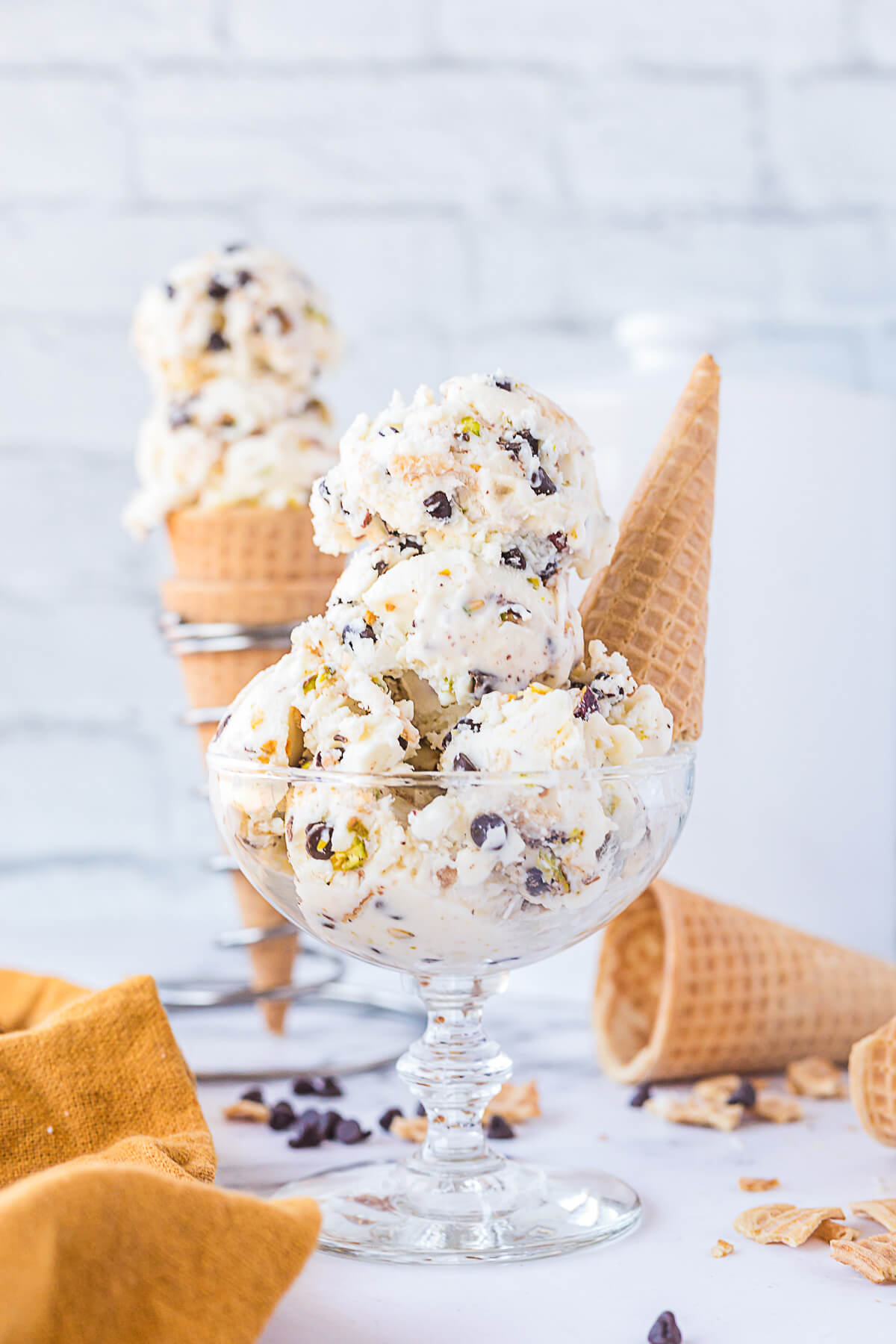 A glass bowl filled with white vanilla ice cream dotted with chocolate chips, pistachios, and broken sugar cone bits.