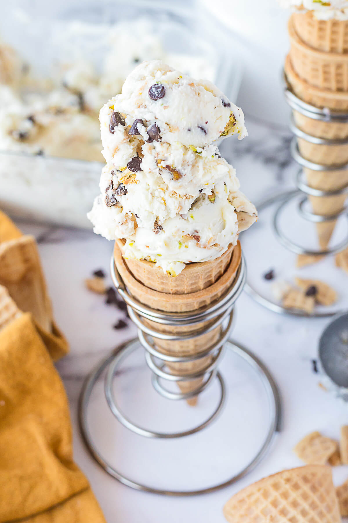 A sugar cone filled with two scoops of white vanilla ice cream dotted with chocolate chips, pistachios, and broken sugar cone bits.