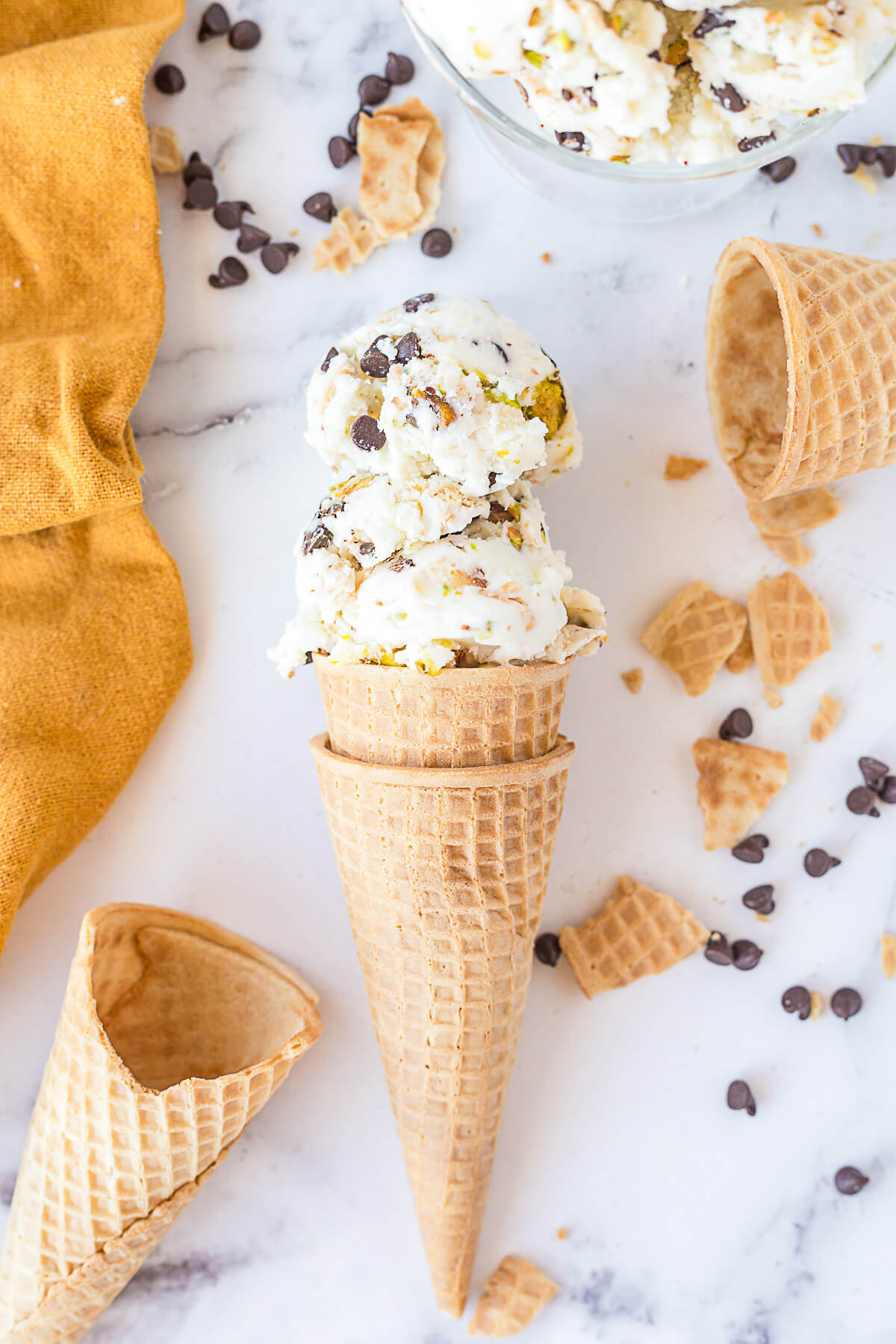 Two sugar cones filled with two scoops of white vanilla ice cream dotted with chocolate chips, pistachios, and broken sugar cone bits.