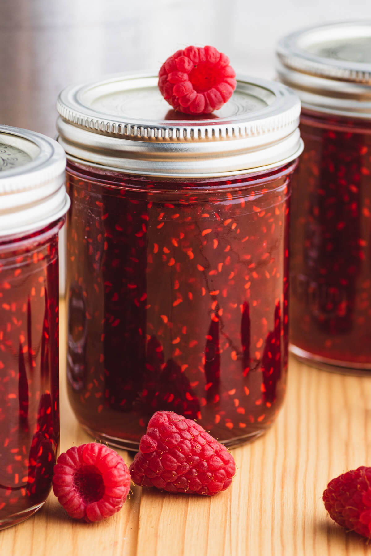 A fresh raspberry sits on top of one jar of raspberry jam in a group of three jars.
