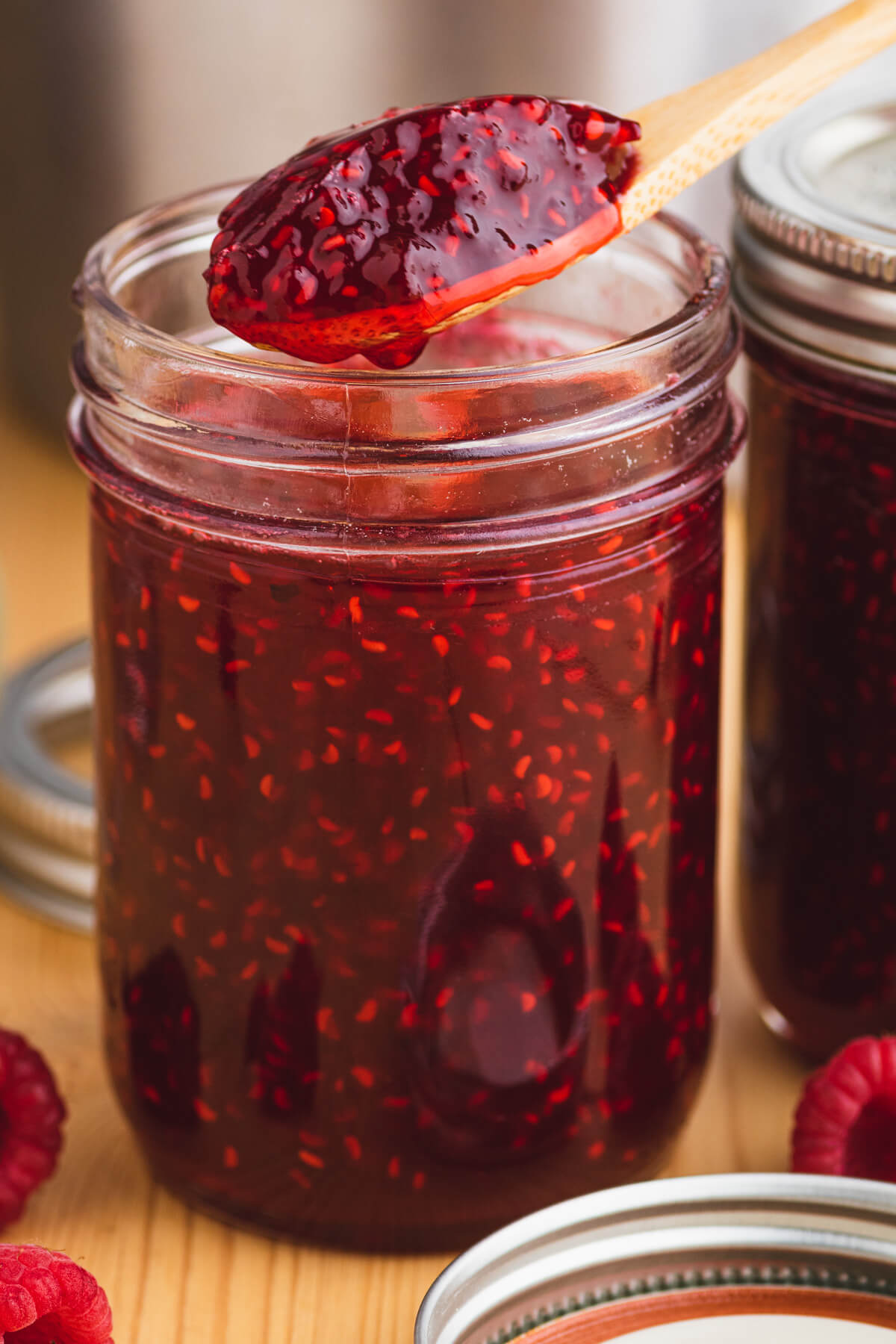 A wooden spoon holds a spoonful of raspberry jam over a jar of the same jam.