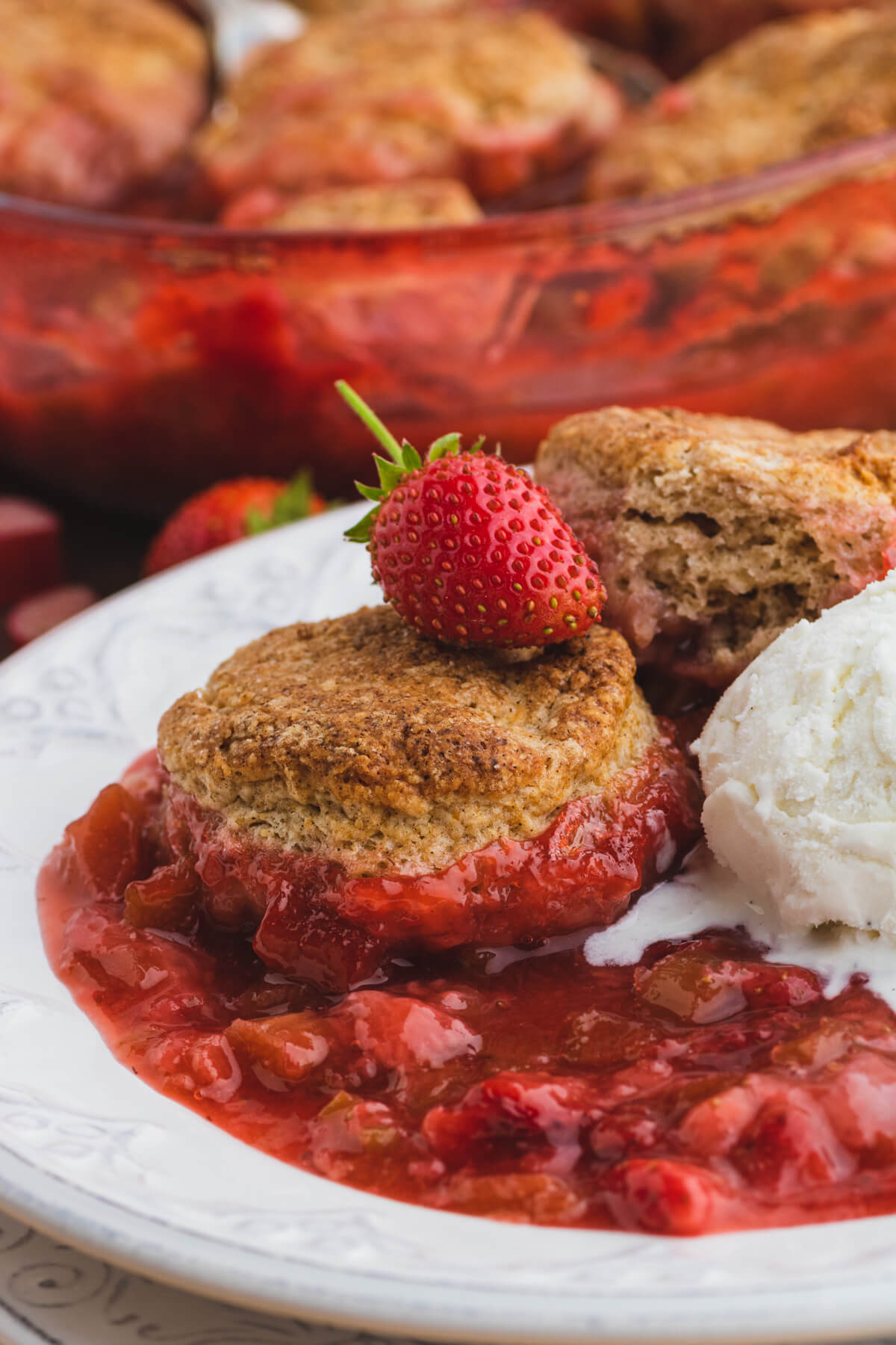 Strawberry Rhubarb Cobbler served on a white plate with a melting scoop of vanilla ice cream and a fresh ripe strawberry.