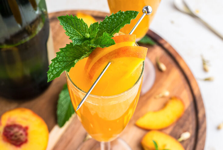 One peach bellini cocktail in a champagne glass garnished with a spring of fresh mint and sliced peaches.