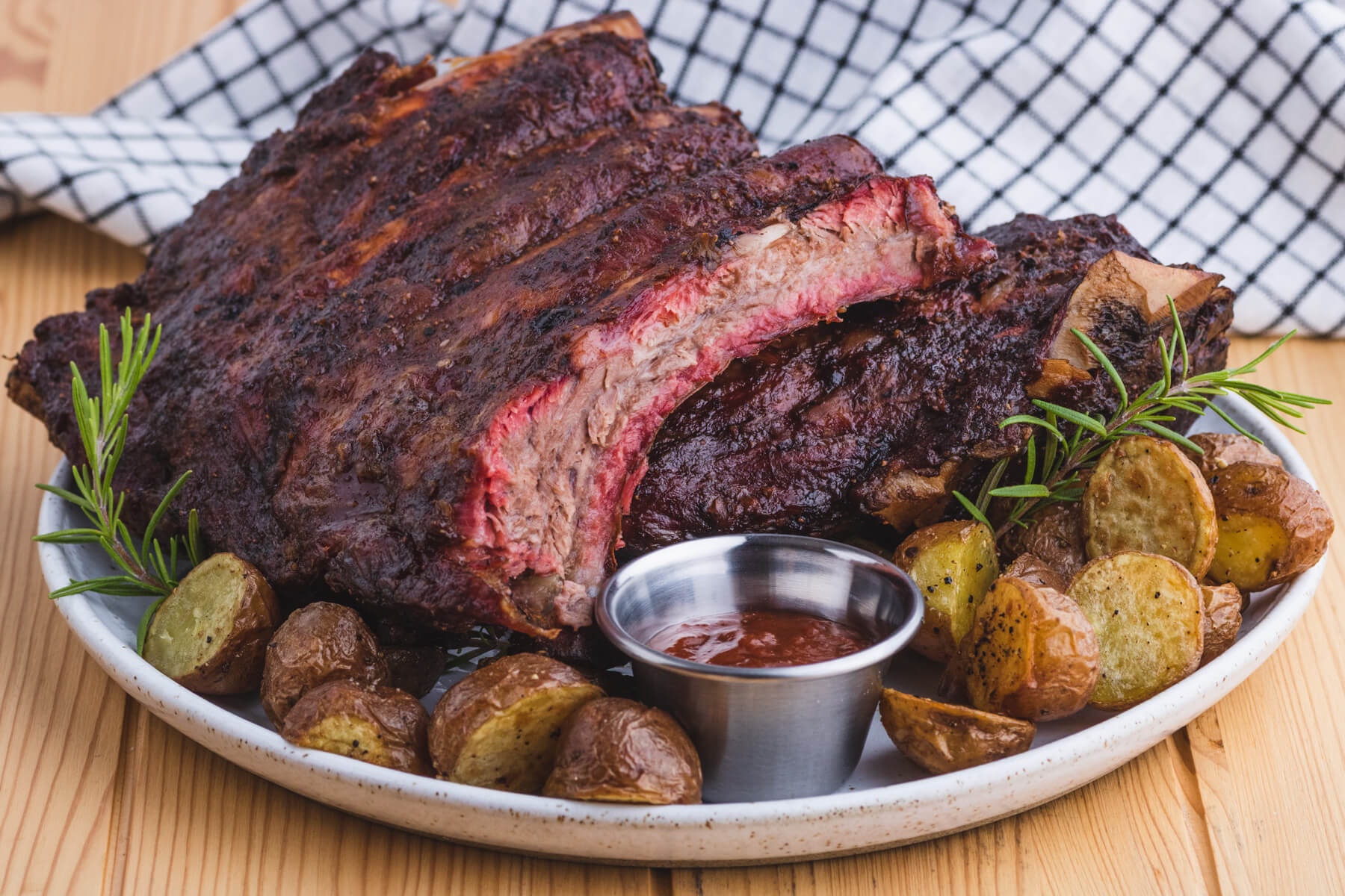 A platter holding a cut rack of cut smoked beef ribs, roasted potatoes, and barbecue sauce.