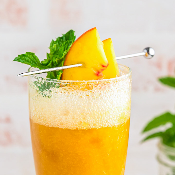 A champagne flute filled with peach puree and bubbly prosecco garnished with fresh peach slices and sprig of mint.