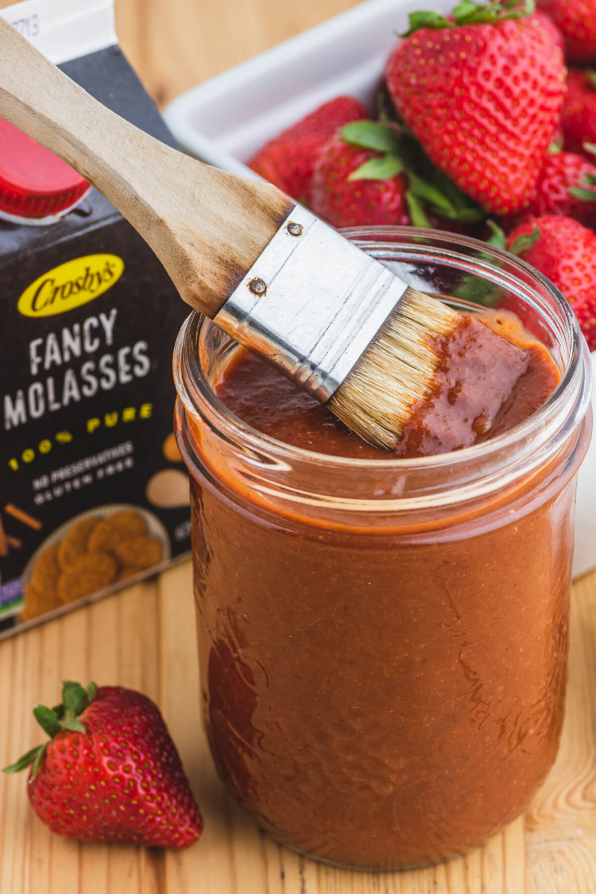 A brush sits in a jar of barbecue sauce surrounded by strawberries, garlic and a package of fancy molasses.