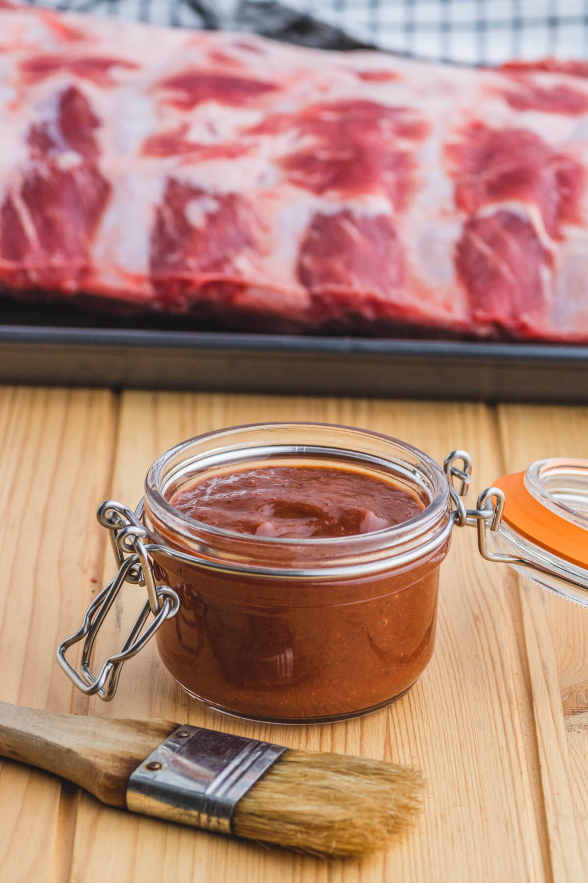A jar of barbecue sauce sits in front of a raw rack of beef ribs.