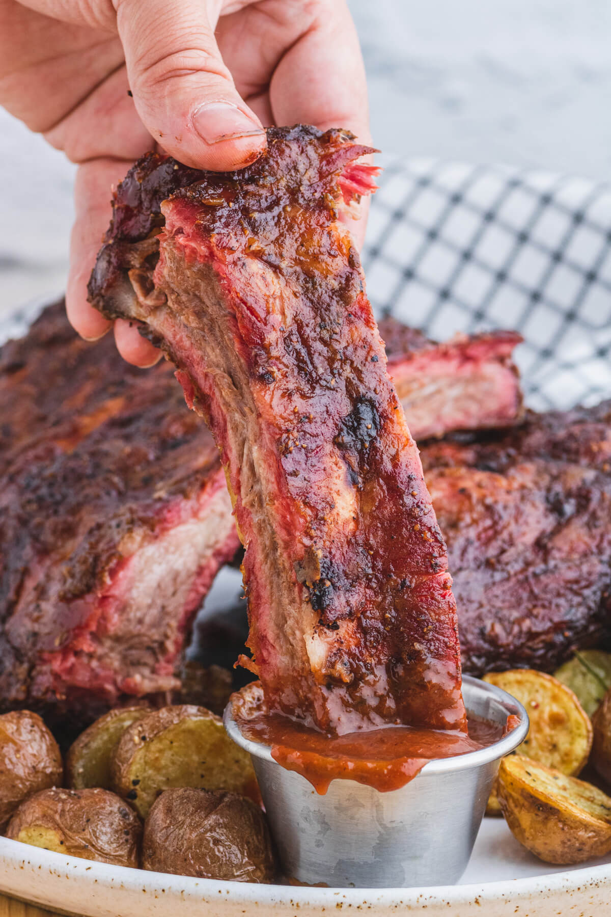 A hand dipping a smoked beef rib into a stainless steel dipping container filled with barbecue sauce.